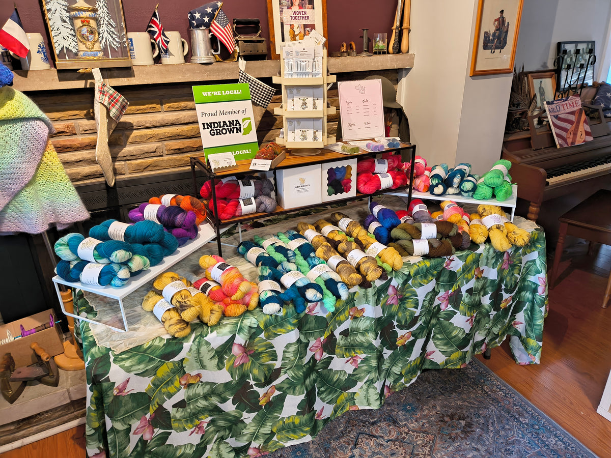 A display table covered with a tropical leaf print cloth, covered in colorful skeins of yarn and fiber from Cat & Rabbit. There are also tools and a sign of the Indiana Grown displayed. 